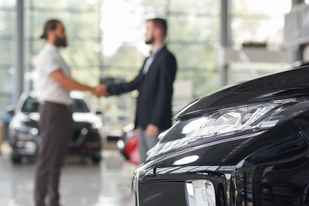 Automobile headlight and men standing behind. Car dealer and client of car dealership shaking hands. Manager and customer making deal on purchasing vehicle. Modern auto showroom.